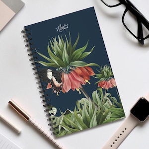 Peut inclure: Cahier à spirales bleu foncé avec une illustration botanique d'une fleur et d'un papillon. Le mot "Notes" est écrit en cursive. Un stylo, des lunettes et une montre connectée sont également visibles.