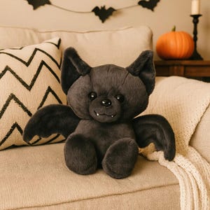 Puede incluir: Un peluche de murciélago gris oscuro con orejas grandes, alas y una cara sonriente. El murciélago está sentado en un sofá beige con un cojín decorativo. Una pequeña calabaza y decoraciones de murciélagos están en el fondo.