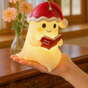 May include: A glowing Christmas-themed night light shaped like a cartoon character, wearing a red Santa hat with a small bell. The character is holding a red book with the words "Merry Christmas". The light is held in a hand.