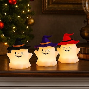 Può includere: Tre luci di Halloween a forma di fantasma luminoso con cappelli da strega neri, viola e rossi. Ogni luce ha una faccia sorridente con occhi e bocca cuciti. Le luci sono su una superficie di legno con un albero di Natale sullo sfondo.