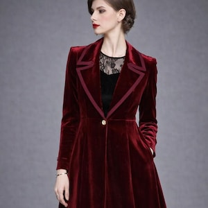 Puede incluir: Abrigo largo de terciopelo burdeos con cuello a medida y cierre de un solo botón dorado. El abrigo presenta un ribete rojo oscuro en contraste y una falda acampanada. Se ve una blusa de encaje negro debajo.
