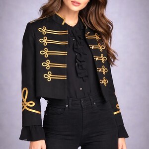 Peut inclure: Veste noire courte avec garniture dorée et détails décoratifs. La veste présente un col montant et des ornements dorés sur le devant et les manches. Le modèle porte un chemisier noir et un jean noir.