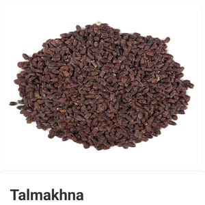 May include: A pile of dark brown Talmakhna seeds against a white background. The seeds are small, oblong, and densely packed together. The image is well-lit, highlighting the texture and color of the seeds.