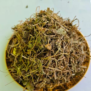 May include: A golden bowl filled with dried herbs and leaves. The herbs are a mix of green and brown, with a tangled, natural appearance. The bowl sits on a white surface, creating a contrast with the earthy tones of the herbs.