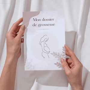 May include: A white card with the text "Mon dossier de grossesse" and a line drawing of a pregnant person. The card has a light pink and beige color scheme with floral accents. The card is held by two hands.