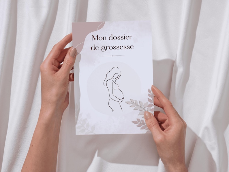 May include: A white card with the text "Mon dossier de grossesse" and a line drawing of a pregnant person. The card has a light pink and beige color scheme with floral accents. The card is held by two hands.