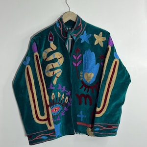 Op de afbeelding: Teal fluwelen jas met geborduurde gouden, bordeauxrode, blauwe en paarse ontwerpen. Met slangen-, hand-, oog- en stermotieven. De jas heeft een hoge kraag en hangt aan een houten hanger.