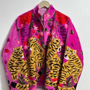 Puede incluir: Chaqueta de terciopelo rosa vibrante con diseños bordados de tigres y flores. La chaqueta presenta cuatro tigres dorados con rayas negras, rodeados de flores rojas, rosas y moradas. Tiene un cuello alto y un forro a rayas.