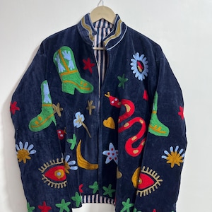 May include: Navy blue jacket with colourful embroidered designs. Features include green cowboy boots, a red snake, flowers, and stars. The jacket has a striped lining and a zip-up front.