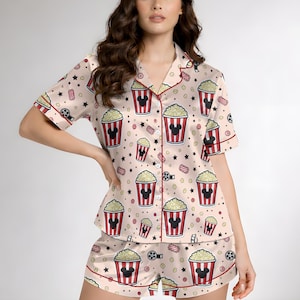 May include: A light pink pajama set with a pattern of popcorn buckets with Mickey Mouse silhouettes, film reels, and stars. The short-sleeved top and shorts have red trim. The set is made of a silky material.