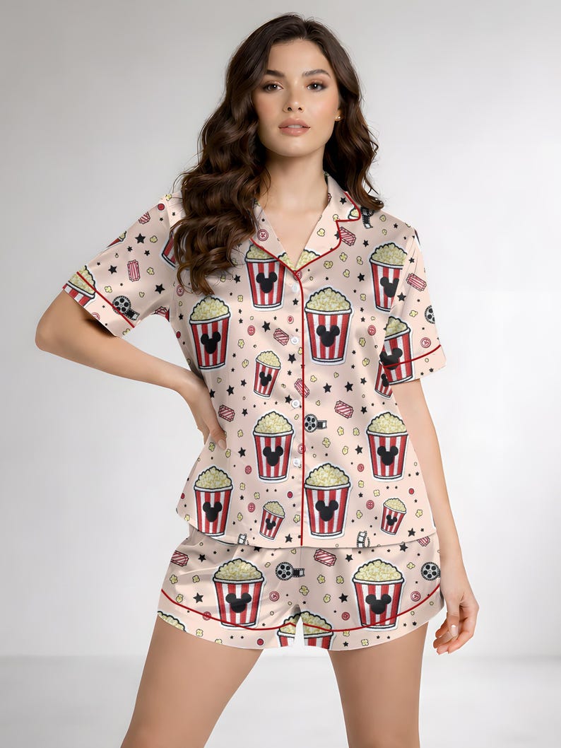 May include: A light pink pajama set with a pattern of popcorn buckets with Mickey Mouse silhouettes, film reels, and stars. The short-sleeved top and shorts have red trim. The set is made of a silky material.