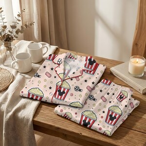 May include: A light pink pajama set with a popcorn and film theme. The set includes a button-down top and trousers, both decorated with popcorn buckets, film reels, and Mickey Mouse silhouettes. The trim is red.
