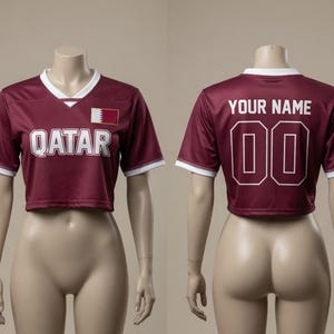 以下が含まれることがあります： 白のトリムと白いブロック文字で「QATAR」という言葉が入った、マルーンのクロップドサッカーユニフォーム。ユニフォームにはカタールの国旗パッチがあり、背面には数字「00」の上に「YOUR NAME」と書かれています。