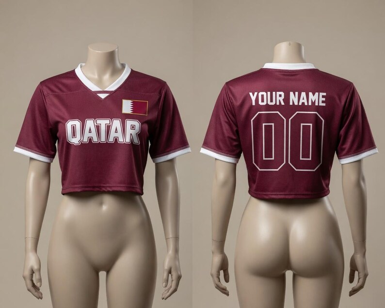 以下が含まれることがあります： 白のトリムと白いブロック文字で「QATAR」という言葉が入った、マルーンのクロップドサッカーユニフォーム。ユニフォームにはカタールの国旗パッチがあり、背面には数字「00」の上に「YOUR NAME」と書かれています。