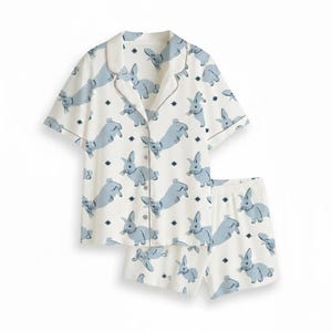 Op de afbeelding: Een tweedelige pyjamaset met een witte achtergrond en een patroon van lichtblauwe konijnen en kleine donkerblauwe sterren. De set bevat een shirt met korte mouwen en bijpassende shorts.