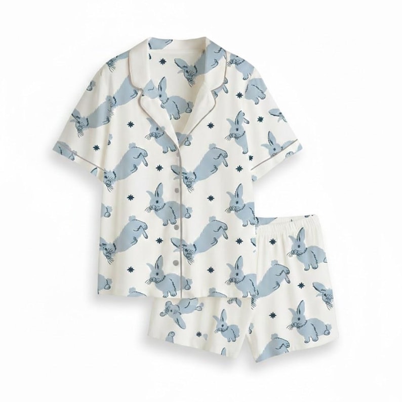 Op de afbeelding: Een tweedelige pyjamaset met een witte achtergrond en een patroon van lichtblauwe konijnen en kleine donkerblauwe sterren. De set bevat een shirt met korte mouwen en bijpassende shorts.