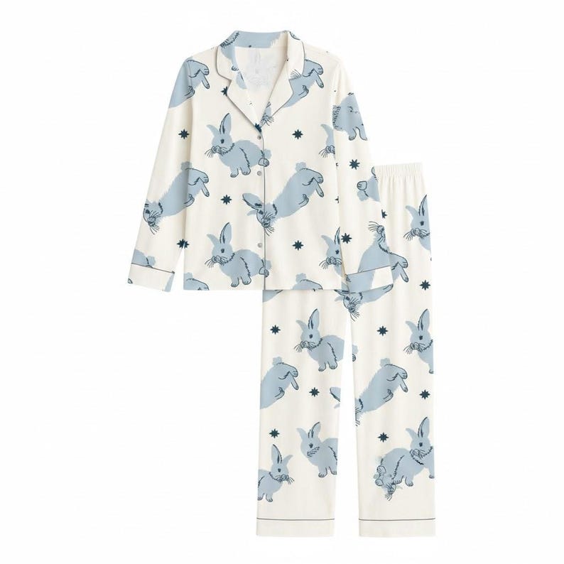 Op de afbeelding: Een tweedelige pyjamaset met een witte achtergrond en een herhaald patroon van lichtblauwe konijnen en donkerblauwe sterren. De set bevat een overhemd met knopen, reverskraag en lange mouwen en een bijpassende broek.