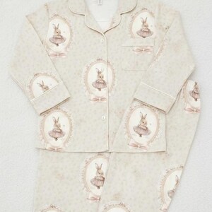 K&ouml;nnte beinhalten: Hellgr&uuml;nes Pyjama-Set mit einem sich wiederholenden Muster von gerahmten Hasenillustrationen in rosa Tutus. Das Set beinhaltet ein Hemd mit Kn&ouml;pfen und Kragen sowie eine passende Hose. Der Pyjama hat wei&szlig;e Paspeln und einen zarten floralen Hintergrund.