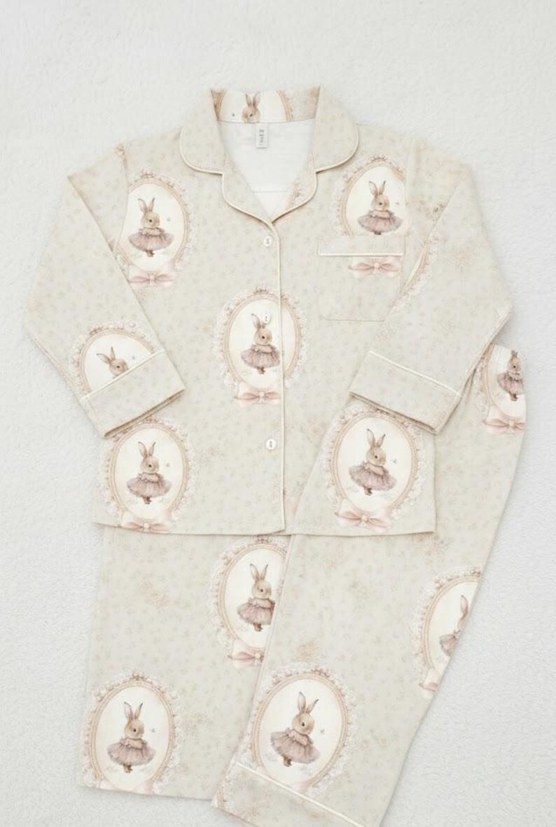 K&ouml;nnte beinhalten: Hellgr&uuml;nes Pyjama-Set mit einem sich wiederholenden Muster von gerahmten Hasenillustrationen in rosa Tutus. Das Set beinhaltet ein Hemd mit Kn&ouml;pfen und Kragen sowie eine passende Hose. Der Pyjama hat wei&szlig;e Paspeln und einen zarten floralen Hintergrund.