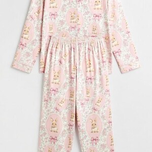 Puede incluir: Conjunto de pijama rosa con un estampado repetido de conejitos en marcos ovalados con lazos rosas y detalles florales. La parte superior de manga larga tiene una botonadura y un cuello con solapas. Los pantalones tienen cintura el&aacute;stica.