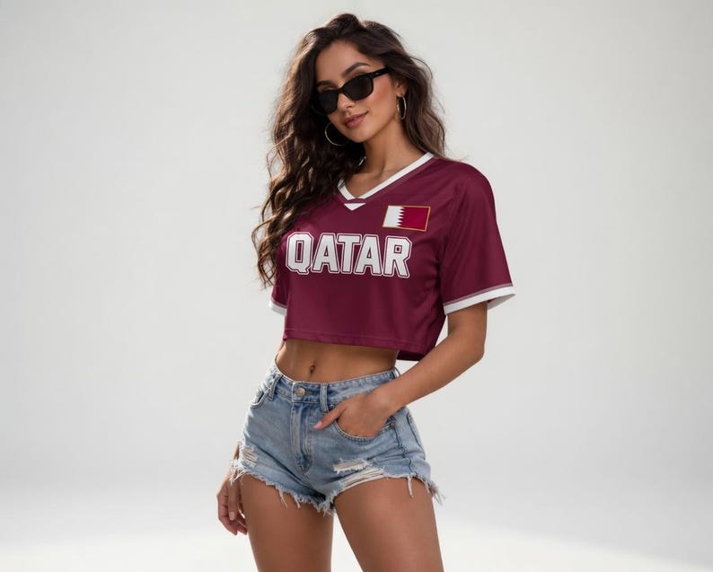 以下が含まれることがあります： 白いトリムと白いブロック文字で「QATAR」という文字が入った、マルーンのクロップトップ。トップにはカタールの国旗パッチとVネックのデザインがあります。モデルはデニムショーツとサングラスを着用しています。