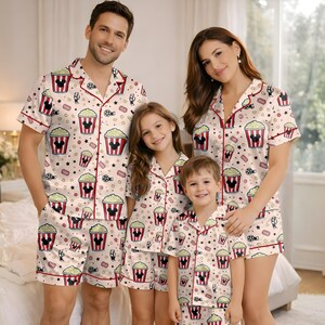 May include: Cream-coloured family pyjama sets featuring a pattern of popcorn buckets, film reels, and Mickey Mouse silhouettes. The pyjamas have red trim and are worn by a family of four. The sets include short-sleeved button-up shirts and shorts.