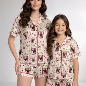May include: Coordinating pyjama sets with a popcorn and Mickey Mouse design. The sets comprise short-sleeved, button-up shirts and matching shorts. The pyjamas are light pink with red and black details. The design includes popcorn buckets, stars, and film reels.