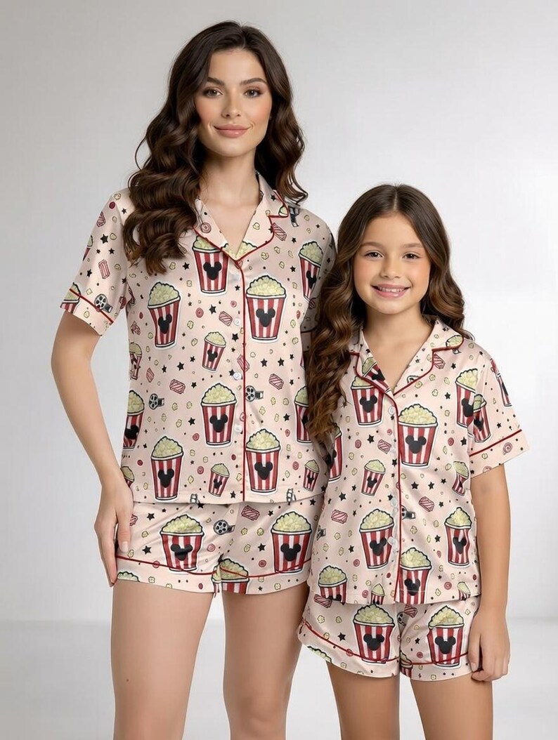 May include: Coordinating pyjama sets with a popcorn and Mickey Mouse design. The sets comprise short-sleeved, button-up shirts and matching shorts. The pyjamas are light pink with red and black details. The design includes popcorn buckets, stars, and film reels.