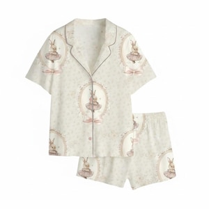 K&ouml;nnte beinhalten: Ein zweiteiliges hellbeige Pyjama-Set mit einem sich wiederholenden Muster eines H&auml;schens in einem Tutu in einem ovalen Rahmen. Das Set beinhaltet ein Hemd mit Kn&ouml;pfen und kurzen &Auml;rmeln sowie passende Shorts. Die Pyjamas haben rosa Akzente.