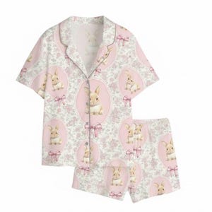 Puede incluir: Un conjunto de pijama de dos piezas con una camisa abotonada de manga corta y pantalones cortos a juego. El conjunto es blanco con un estampado floral rosa y medallones ovalados rosas con un conejito y lazos rosas. Hecho de tela ligera.
