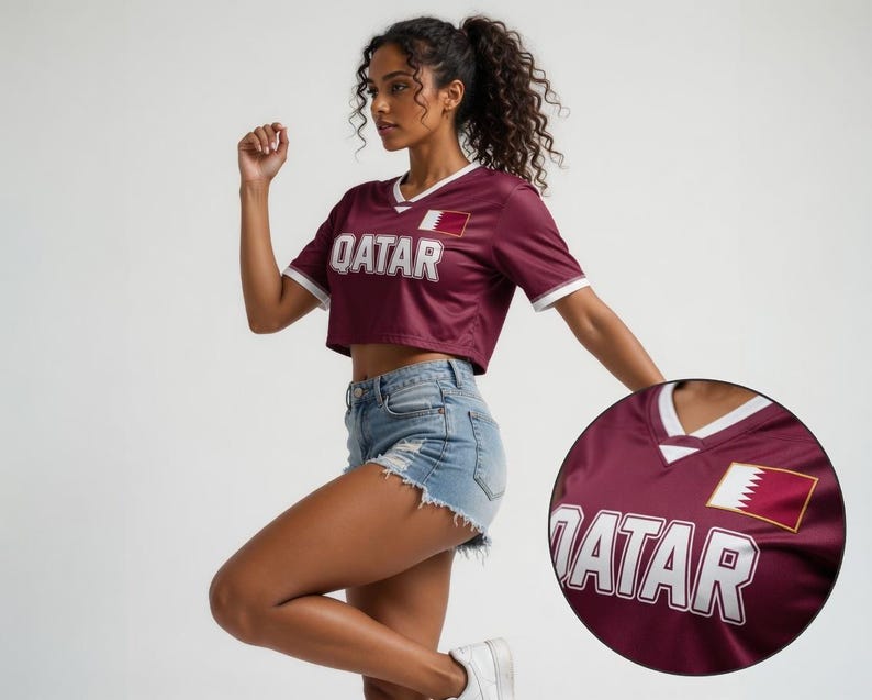 以下が含まれることがあります： 白のトリムと白いブロック文字で「QATAR」という文字が入った、マルーンのクロップトップジャージー。ジャージーの胸にはカタールの国旗のパッチが付いています。モデルはデニムショーツと白いスニーカーを着用しています。