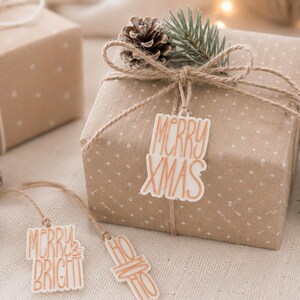 Drei Geschenkanhänger Weihnachten in Creme und Kupfer an und neben einem natürlich verpackten Weihnachtsgeschenk mit Tannenzweig und Zapfen.