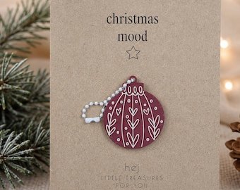 Könnte beinhalten: Ein kastanienbrauner Weihnachtsbaumschmuck-Anhänger mit weißen Linien-Details an einer weißen Kugelkette. Der Anhänger ist auf einer rechteckigen beigen Karte befestigt, die den Text "christmas mood" mit einem Sternsymbol und "hej LITTLE TREASURES FOR YOU" zeigt.