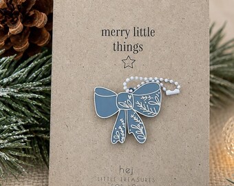 Könnte beinhalten: Ein blauer schleifenförmiger Anhänger mit weißen floralen Linienmustern, befestigt an einer weißen Kugelkette. Die Schleife ist auf einer rechteckigen beigen Karte montiert, die den Text merry little things, einen Stern und hej little treasures for you zeigt.