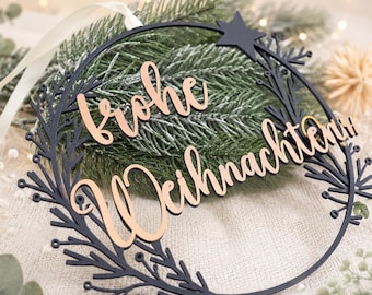 Könnte beinhalten: Ein kreisförmiger schwarzer Anhänger mit einem Ast-Design und einem Stern an der Oberseite. Der Schriftzug "frohe Weihnachten" ist in einer metallisch roségoldenen Schreibschrift in der Mitte angebracht. Ein weißes Band dient zum Aufhängen.