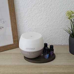 Könnte beinhalten: Ein weiß-brauner ätherischer Öl Diffusor der Marke "doTERRA" steht auf einer Holzoberfläche. Zwei kleine ätherische Ölflaschen befinden sich neben dem Diffusor. Ein gerahmter Druck mit dem Text "collect moments not things" befindet sich links.