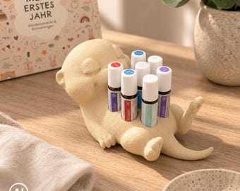 May include: A cream-colored, textured otter-shaped figurine serving as a holder for six small cylindrical essential oil bottles with colorful caps. The otter is lying on its back with its eyes closed, and the bottles are inserted into its torso.