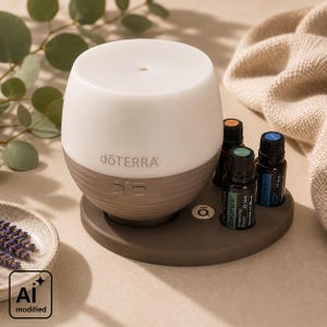 Könnte beinhalten: Ein dōTERRA Diffusor für ätherische Öle mit weißem Oberteil und geripptem grauen Sockel, ausgestattet mit Tasten für Nebel- und Lichtsteuerung. Drei kleine dunkle Glasflaschen mit ätherischen Ölen stehen auf dem runden grauen Sockel.