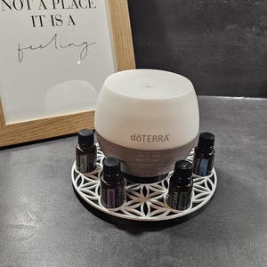 Könnte beinhalten: Ein weiß-grauer Diffusor für ätherische Öle mit dem Markennamen "dōTERRA". Der Diffusor steht auf einem weißen Dekotablett, umgeben von kleinen ätherischen Ölflaschen. Im Hintergrund ein gerahmter Druck mit dem Text "HOME IS NOT A PLACE IT IS A feeling".