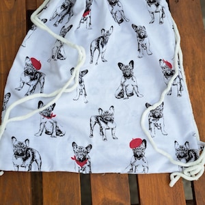 Könnte beinhalten: Weißer Turnbeutel mit einem sich wiederholenden Muster aus schwarz-weißen Illustrationen von Französischen Bulldoggen. Einige Bulldoggen tragen rote Baretts oder Bandanas. Die Tasche hat einen weißen Kordelzugverschluss und wird auf einer Holzoberfläche präsentiert.