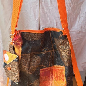 May include: A fabric shoulder bag featuring a dark brown and black leaf pattern with orange accents. The bag has a bright orange zip closure and matching orange nylon straps. A small, detachable matching pouch is clipped to the side strap.