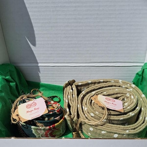 May include: Two coiled fabric bag straps with metal hardware inside a white box. One strap features a dark green floral pattern, and the other is a solid olive green with white polka dots. Each strap has a light pink tag labelled Mager Bags.