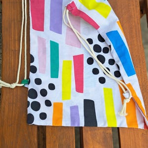 May include: Two drawstring bags with a white background and a colourful abstract pattern of vertical rectangles and black polka dots. The bags have beige drawstrings and are resting on a wooden surface.