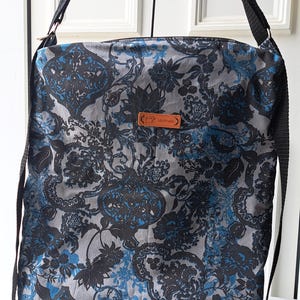 May include: A square crossbody bag featuring a grey fabric base with a dark blue and black floral damask pattern. The bag includes a black adjustable strap and a small rectangular brown leather tag centred on the front with the word handmade.