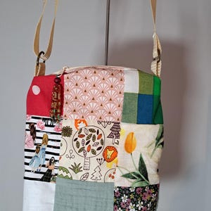 Op de afbeelding: Een patchwork crossbodytas met een beige band. De tas heeft een verscheidenheid aan stofpatronen, waaronder bloemen-, geometrische en geïllustreerde ontwerpen. De tas heeft een ritssluiting en een decoratieve kwast.