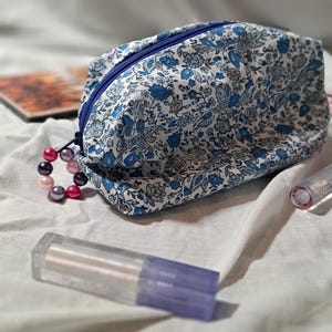 May include: A floral print cosmetic bag with a blue zipper and a beaded pull. The bag features a pattern of blue flowers on a white background. Two lip gloss tubes are in the foreground.