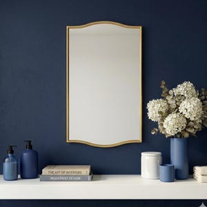 May include: A gold-framed mirror with a curved top hangs above a white shelf. The shelf holds blue soap dispensers, books, a white jar, a blue vase with white flowers, and a blue cup. The background is a dark blue wall.