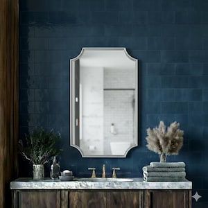 Può includere: Uno specchio argentato con cornice ad arco è appeso sopra un mobile da bagno in legno scuro. Il mobile ha un piano in marmo, rubinetti in ottone e una pila di asciugamani piegati. Lo sfondo è una parete piastrellata blu scuro.