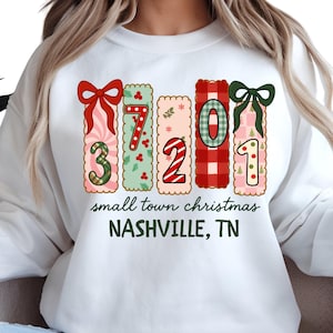May include: White sweatshirt with a colorful Christmas-themed design. The design includes the numbers 37201 in decorative rectangles, bows, and the text "small town christmas NASHVILLE, TN". The numbers are in various patterns and colors.