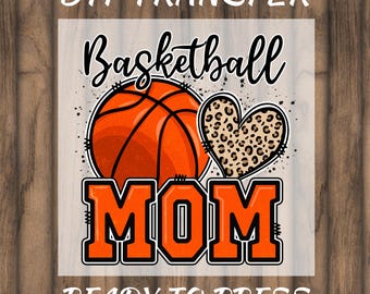 Puede incluir: Diseño de transferencia DTF con una pelota de baloncesto y un corazón con estampado de leopardo. La palabra "Basketball" está escrita encima de la pelota, y "MOM" en letras naranjas. La frase "READY TO PRESS" está en la parte inferior.